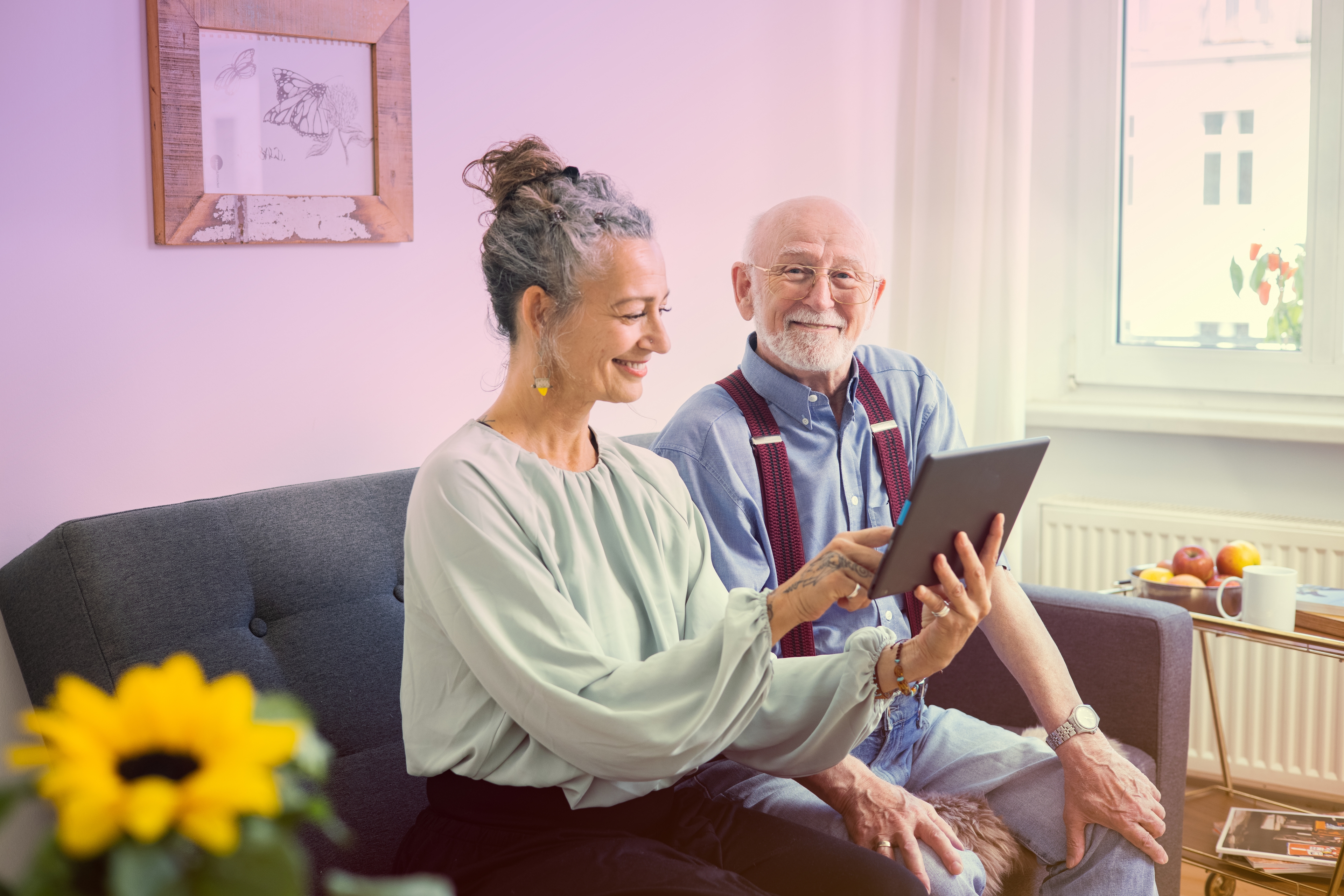 Ein glückliches älteres Ehepaar, das gemeinsam auf dem Sofa auf das Tablet schaut, während der Mann in unsere Richtung schaut und lächelt.